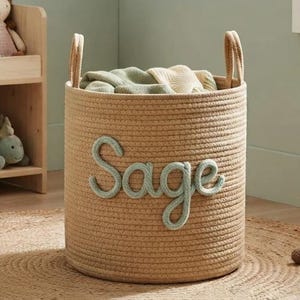 May include: A cylindrical woven rope storage basket in a natural tan color featuring the name Sage in raised, sage green cursive lettering on the front. The basket contains folded light-colored fabric and has two sturdy handles on the top rim.