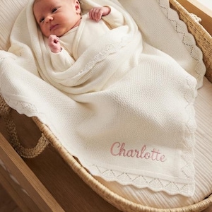 May include: A woven wicker bassinet containing a baby wrapped in a cream-colored knit blanket. The blanket features a delicate scalloped edge and the name Charlotte embroidered in soft pink thread. A small plush rabbit toy sits nearby.