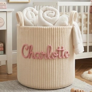 May include: A beige corduroy storage basket with the name "Charlotte" in pink script. The basket is filled with rolled white blankets and towels. The nursery setting includes a crib, artwork, and a mobile.