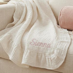 May include: A cream-colored baby blanket with a ruffled edge and the name "Sienna" embroidered in pink. The blanket is draped over a beige sofa, next to a pink bolster pillow and a wooden rattle.