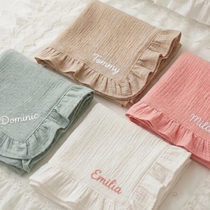 May include: Four folded muslin blankets with ruffled edges in sage green, beige, pink, and white. Each blanket features white or coral embroidered names: Dominic, Tommy, Millie, and Emilia, stitched in a cursive script on the fabric surface.