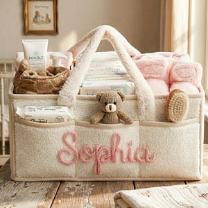 Puede incluir: Un organizador de bebé color crema con el nombre "Sophia" bordado en rosa. El organizador tiene múltiples bolsillos, un osito de peluche y un cepillo. Contiene artículos esenciales para bebés como pañales y una manta rosa. El organizador tiene una textura suave y afelpada.