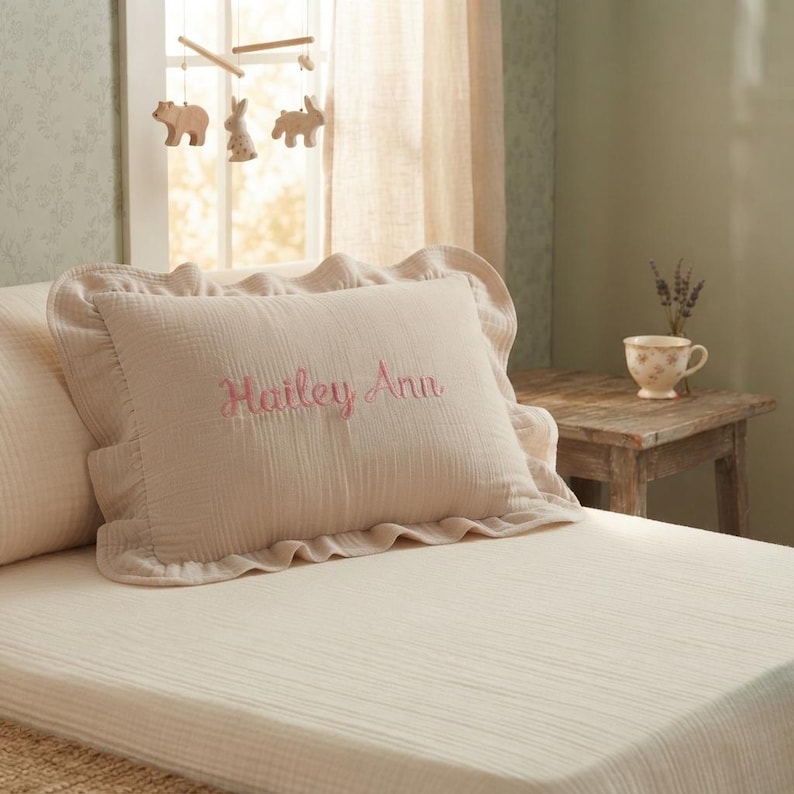 May include: A rectangular beige pillowcase with a ruffled edge, featuring the embroidered name Hailey Ann in pink script lettering. The fabric has a textured, crinkled appearance, and the pillow is positioned on a matching beige bed sheet.