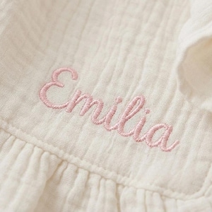 May include: A cream-colored muslin fabric garment featuring the name Emilia embroidered in light pink cursive thread. The textile has a distinct crinkled, textured surface with visible vertical stitching and a ruffled edge detail.