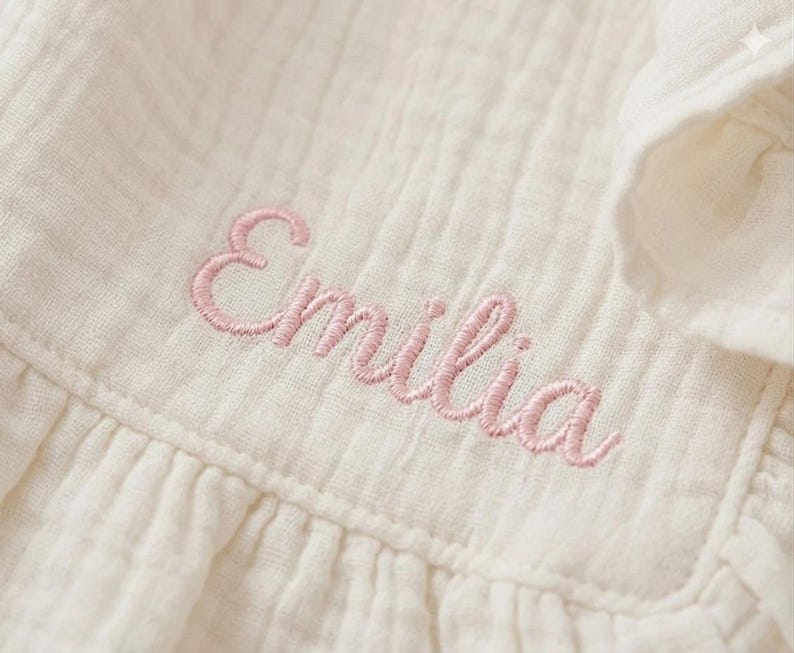 May include: A cream-colored muslin fabric garment featuring the name Emilia embroidered in light pink cursive thread. The textile has a distinct crinkled, textured surface with visible vertical stitching and a ruffled edge detail.