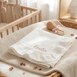 May include: A white muslin baby blanket with a ruffled edge and the name Emilia embroidered in pink thread. The blanket is folded and placed on a textured beige surface next to a small woven rattan rattle with a light pink ribbon bow attached.