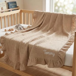 May include: A light tan muslin baby blanket with a ruffled border and the name Tommy embroidered in white script. The blanket is draped over a wooden crib, featuring a textured fabric finish and a soft, neutral color palette throughout the design.