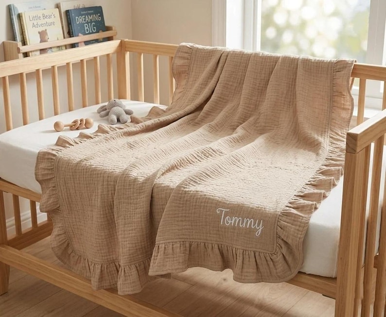 May include: A light tan muslin baby blanket with a ruffled border and the name Tommy embroidered in white script. The blanket is draped over a wooden crib, featuring a textured fabric finish and a soft, neutral color palette throughout the design.
