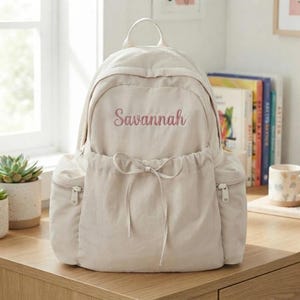 Pode incluir: Uma mochila bege com o nome Savannah bordado em letra cursiva rosa no bolso frontal. A mochila inclui uma alça superior, dois bolsos laterais com fechos prateados e um bolso frontal com um fecho de cordão decorativo.