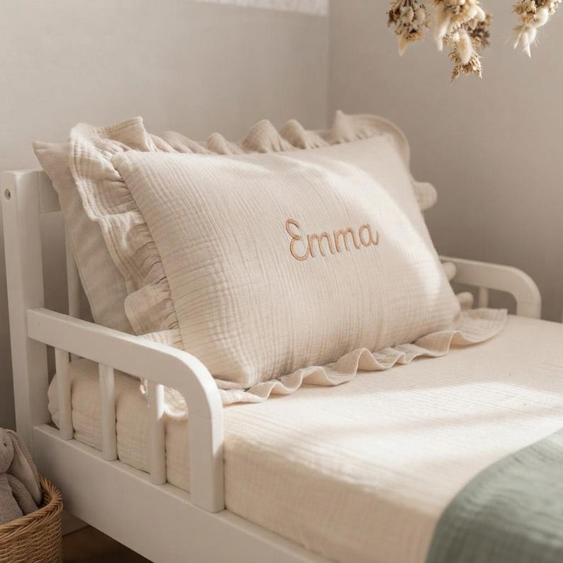 May include: A rectangular cream-colored pillow with a ruffled edge and the name Emma embroidered in a light brown script font on the front. The pillow is made of a textured fabric and is positioned on a white wooden bed frame with a matching sheet.