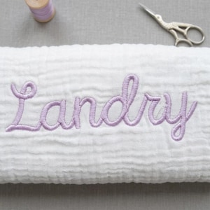 May include: A white muslin fabric blanket featuring the name Landry embroidered in light purple cursive script. The blanket is positioned near a small spool of matching light purple thread and a pair of silver metal embroidery scissors.