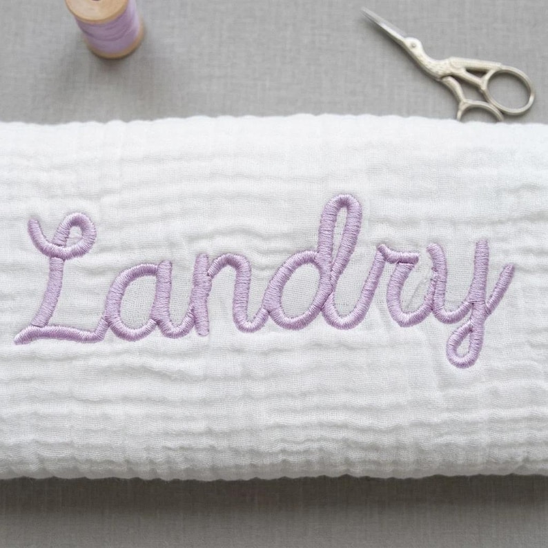 May include: A white muslin fabric blanket featuring the name Landry embroidered in light purple cursive script. The blanket is positioned near a small spool of matching light purple thread and a pair of silver metal embroidery scissors.