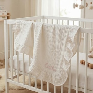 May include: A cream-colored muslin baby blanket draped over a white wooden crib rail. The blanket features a ruffled edge and is embroidered with the name Emilia in light pink script lettering. The fabric has a soft, crinkled, multi-layered texture.