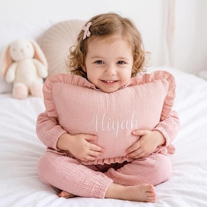 May include: A dusty pink rectangular throw pillow with ruffled edges and the name Aliyah embroidered in white cursive script on the front. The pillow is made of a textured fabric and is held by a child wearing a matching pink outfit.