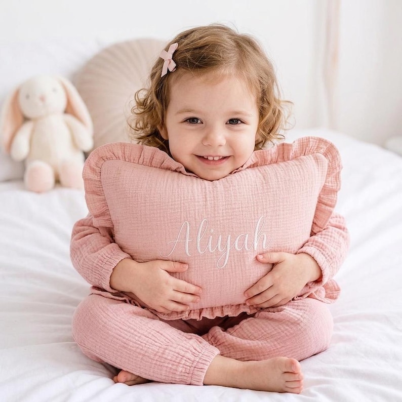 May include: A dusty pink rectangular throw pillow with ruffled edges and the name Aliyah embroidered in white cursive script on the front. The pillow is made of a textured fabric and is held by a child wearing a matching pink outfit.