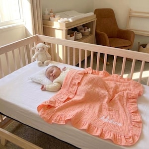 May include: A peach-colored muslin baby blanket with a ruffled edge and the name Adeline embroidered in white script. The blanket is draped over a baby lying on a white sheet in a light-colored wooden crib next to a plush white lamb toy.