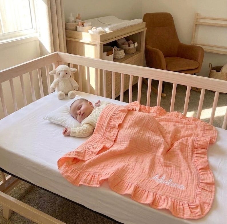 May include: A peach-colored muslin baby blanket with a ruffled edge and the name Adeline embroidered in white script. The blanket is draped over a baby lying on a white sheet in a light-colored wooden crib next to a plush white lamb toy.
