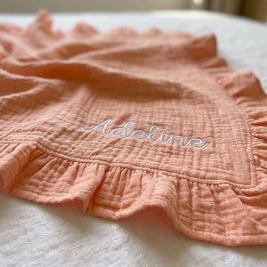 May include: A peach-colored muslin blanket featuring a ruffled border and the name Adeline embroidered in white cursive script. The fabric has a crinkled, textured appearance with soft folds throughout the material, highlighting the stitching.