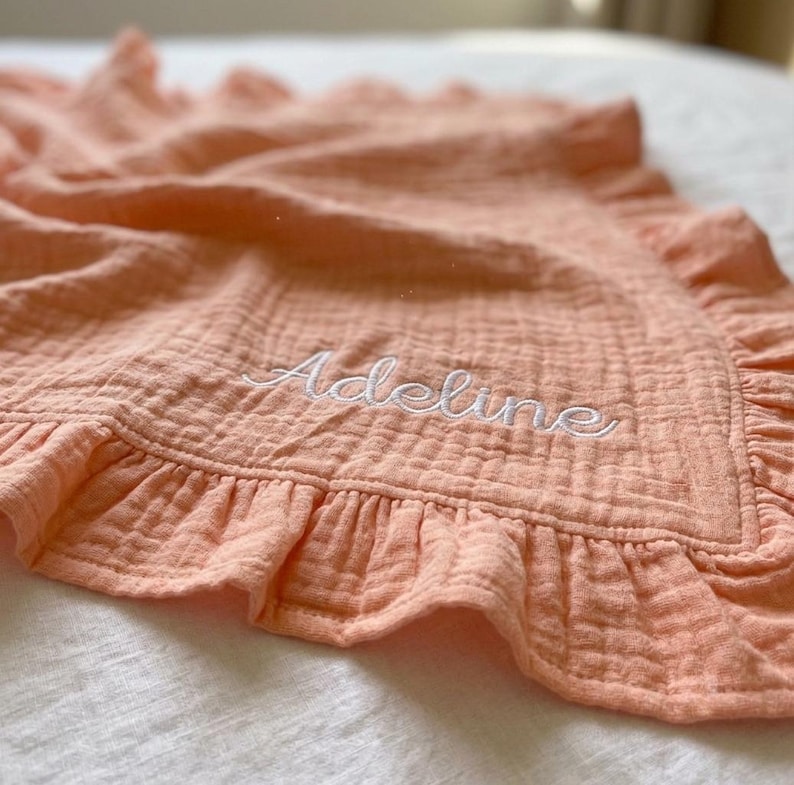 May include: A peach-colored muslin blanket featuring a ruffled border and the name Adeline embroidered in white cursive script. The fabric has a crinkled, textured appearance with soft folds throughout the material, highlighting the stitching.