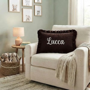 May include: A rectangular dark brown throw pillow with a ruffled edge and the name Lucca embroidered in white script lettering. The pillow is positioned on a light-colored upholstered armchair, featuring a textured fabric surface and a soft appearance.