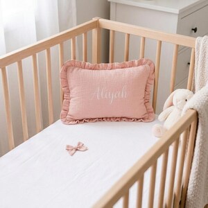May include: A rectangular dusty pink muslin pillow with a ruffled edge and the name Aliyah embroidered in white script lettering. The pillow is placed on a white sheet inside a light-colored wooden crib, accompanied by a small pink fabric bow.