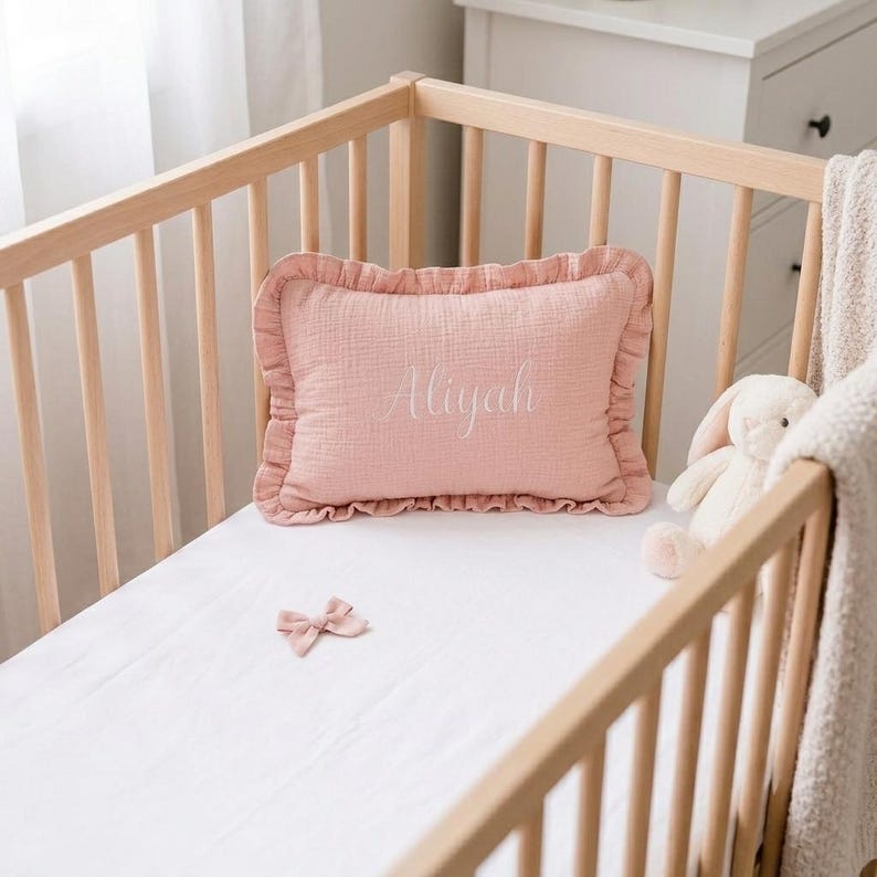 May include: A rectangular dusty pink muslin pillow with a ruffled edge and the name Aliyah embroidered in white script lettering. The pillow is placed on a white sheet inside a light-colored wooden crib, accompanied by a small pink fabric bow.