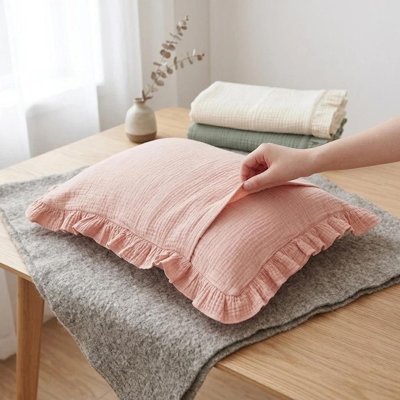 May include: A rectangular pillowcase made of soft, textured pink cotton muslin fabric. The pillowcase features a delicate ruffled edge trim along the perimeter and an envelope-style closure opening on the front side, showcasing the fabric's layered weave.