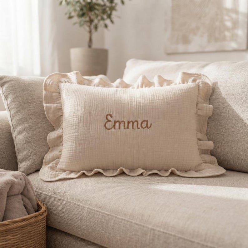 May include: A rectangular beige throw pillow featuring a ruffled border and the name Emma embroidered in brown thread in the center. The pillow is made of a textured fabric and is positioned on a matching beige sofa.