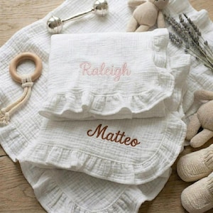 May include: Two white muslin blankets with ruffled edges, each embroidered with a name. The top blanket features the name Raleigh in pink script, and the bottom blanket features the name Matteo in brown script, accompanied by a rattle and plush toys.