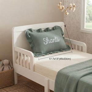 May include: A sage green rectangular pillow with a ruffled edge and the name Shanti embroidered in white script. The pillow measures 17.7 by 13 inches. It is placed on a white toddler bed with a matching sage green blanket and a cream-colored sheet.