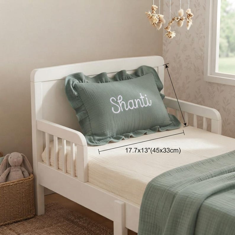 May include: A sage green rectangular pillow with a ruffled edge and the name Shanti embroidered in white script. The pillow measures 17.7 by 13 inches. It is placed on a white toddler bed with a matching sage green blanket and a cream-colored sheet.