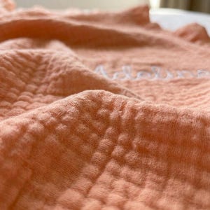 May include: A peach-colored muslin blanket featuring a textured, crinkled grid pattern. The fabric is soft and lightweight, with the name Adeline embroidered in white cursive script across the center of the folded, draped material.