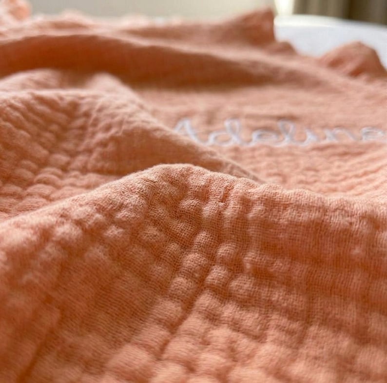 May include: A peach-colored muslin blanket featuring a textured, crinkled grid pattern. The fabric is soft and lightweight, with the name Adeline embroidered in white cursive script across the center of the folded, draped material.