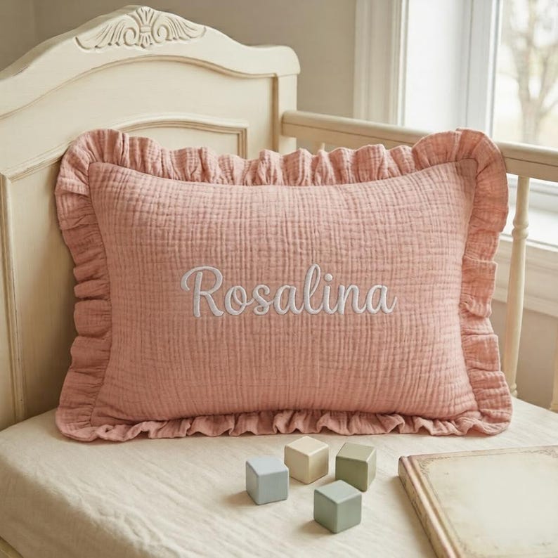May include: A rectangular, dusty pink muslin pillow featuring a ruffled border and the name Rosalina embroidered in white script lettering across the center. The pillow has a textured, crinkled fabric surface and is positioned on a bed.
