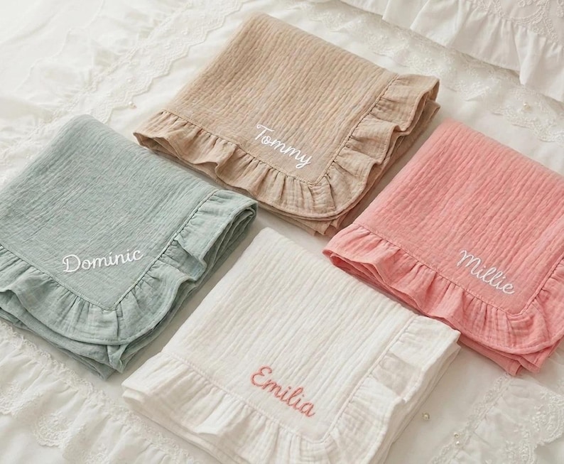 May include: Four folded muslin blankets with ruffled edges in sage green, beige, pink, and white. Each blanket features white or coral embroidered names: Dominic, Tommy, Millie, and Emilia, stitched in a cursive script on the fabric surface.