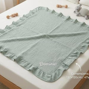 May include: A square sage green muslin baby blanket featuring a ruffled border and the embroidered name Dominic. The blanket includes printed text stating dimensions of 120x120cm and 47x47 inches. The fabric has a crinkled texture throughout the surface.