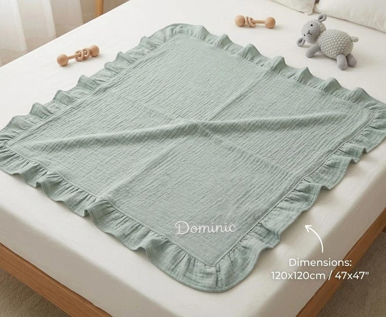 May include: A square sage green muslin baby blanket featuring a ruffled border and the embroidered name Dominic. The blanket includes printed text stating dimensions of 120x120cm and 47x47 inches. The fabric has a crinkled texture throughout the surface.
