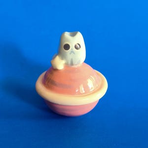 Puede incluir: Un joyero de cerámica con una figura de gato azul claro con grandes ojos negros y una pequeña estrella amarilla a un lado. El gato se asienta sobre un recipiente redondo de color rosa con forma de planeta y un anillo color crema en el centro.