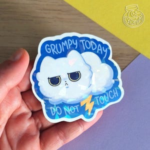 Op de afbeelding: Een holografische sticker met een chagrijnige witte kat en de tekst "GRUMPY TODAY DO NOT TOUCH". De kat staat in een blauwe wolk met een bliksem.