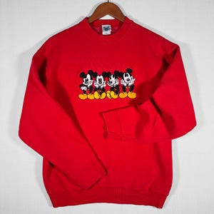 May include: A red crewneck sweatshirt featuring an embroidered design of four Mickey Mouse characters. The cartoon figures are in black, white, and yellow. The sweatshirt is displayed on a wooden hanger.