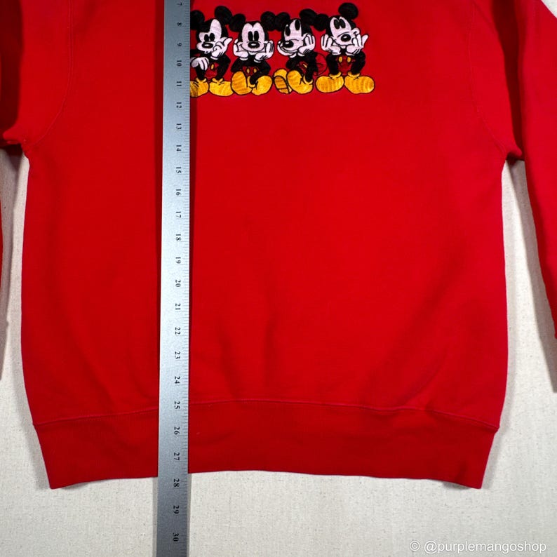 May include: Red sweatshirt featuring an embroidered design of four Mickey Mouse characters. The design includes black, white, and yellow details. The sweatshirt has a crew neck and long sleeves.