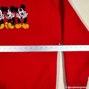 May include: A red sweatshirt featuring an embroidered design of three Mickey Mouse characters. The cartoon characters are in black, yellow, and red. The sweatshirt has a crew neck and long sleeves.