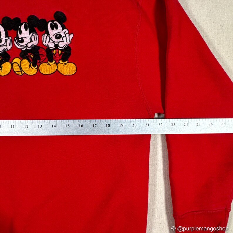 May include: A red sweatshirt featuring an embroidered design of three Mickey Mouse characters. The cartoon characters are in black, yellow, and red. The sweatshirt has a crew neck and long sleeves.