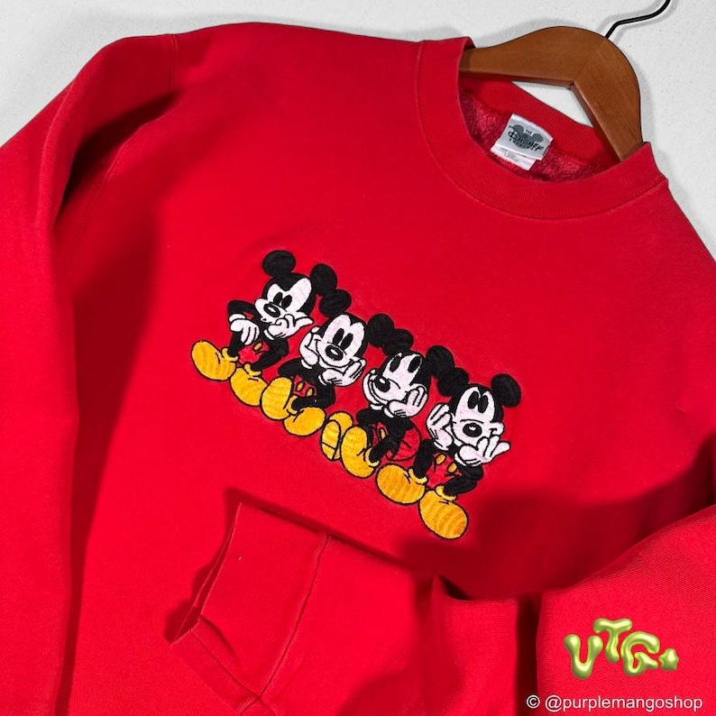 May include: A bright red sweatshirt featuring an embroidered design of four Mickey Mouse characters in a row. The characters are in black, white, red, and yellow. The sweatshirt has a pocket and a crew neck.