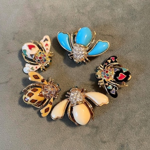 May include: Five decorative bee brooches arranged in a circle. Each brooch has a gold-tone metal design with colourful enamel and rhinestone accents. The brooches feature various wing and body colours, including white, yellow, black, and turquoise.