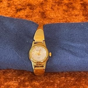 May include: A gold-toned wristwatch with a round face and a gold band. The watch face has a light-colored background with gold hands and markers. The watch is resting on a blue surface.