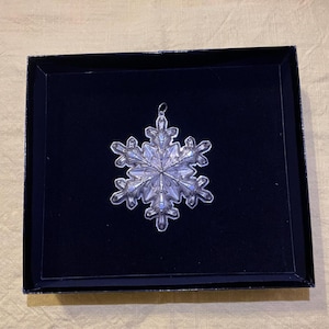 May include: A clear, snowflake-shaped ornament with intricate details, displayed in a black box with a dark blue interior. The ornament has a silver-colored hanger and is likely made of glass or crystal. A festive holiday decoration.