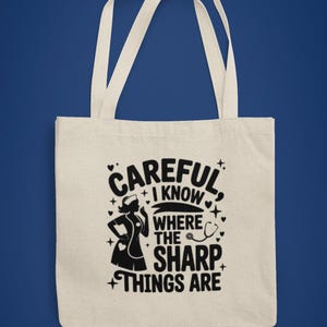 May include: Cream tote bag with white handles. The bag features a black graphic of a nurse and the text "CAREFUL I KNOW WHERE THE SHARP THINGS ARE". The design includes stars and a stethoscope.