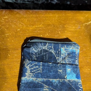 Peut inclure: Petite pochette rectangulaire bleu marine avec un motif de carte en beige et blanc. La pochette a une fermeture éclair noire et est en tissu. Le motif de la carte comprend du texte et des éléments géographiques.