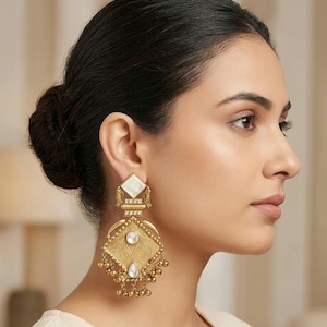 May include: Gold-coloured drop earrings with a square, white stone at the top and a diamond-shaped, textured design below. The earrings feature a central white stone and dangling gold beads. The earrings are worn by a person with dark hair.